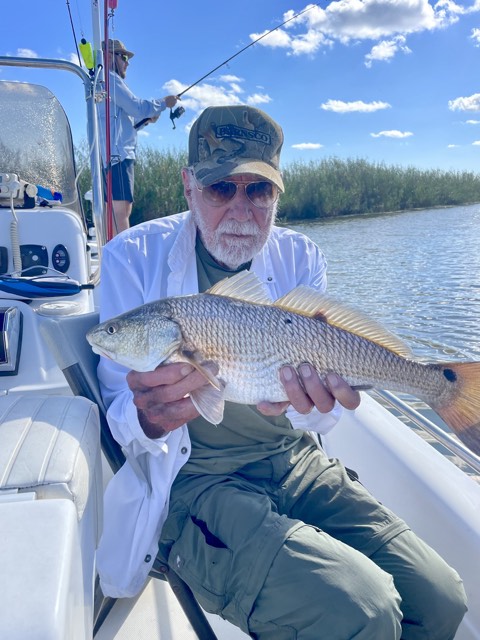 Man on a boat holding a redfish. Another person fishing in the background. Sunny day, water and marsh visible.