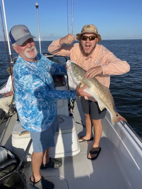 Two people on a boat, holding a large red fish. Happy expressions, blue water, sunny day.