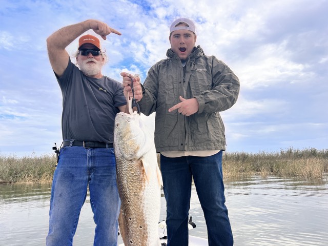 Two people holding a large fish on a boat; cloudy sky background.