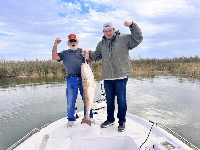 Two men flexing, holding a large fish on a boat. Cloudy sky, marsh background.