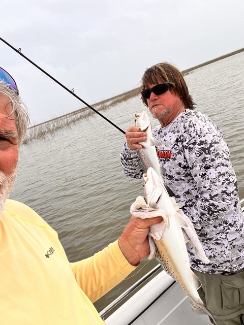 Two men on a boat fishing, holding speckled trout. One man wears sunglasses and a camouflage shirt. Overcast skies.