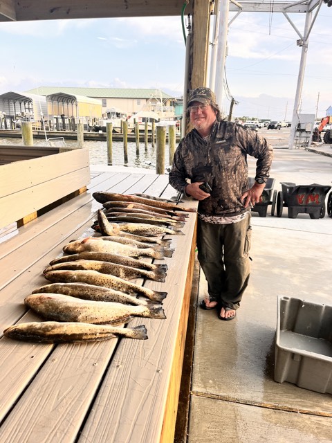 Fisherman stands proudly next to a long wooden table displaying several speckled trout.