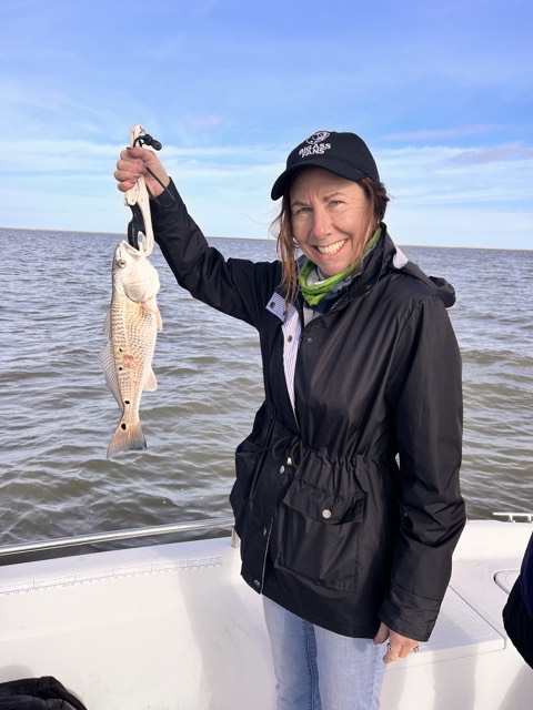 Woman on boat smiling, holding up a fish, ocean in background.