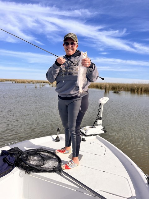 Woman on a boat holding a fish, wearing a hat, hoodie, and pants, on a sunny day.