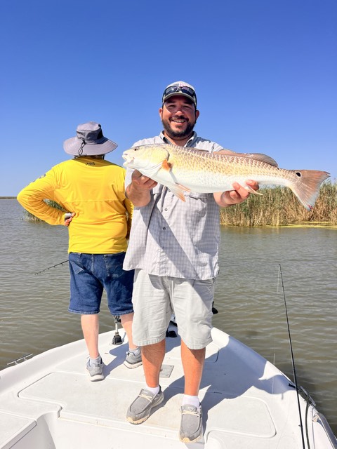 Man holding a redfish on a boat, smiling. Another person watches from behind. Water and reeds in the background.