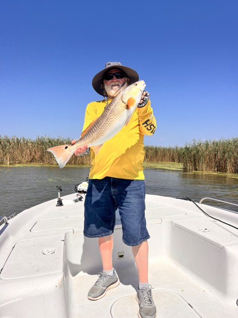 Man on boat holding a large redfish. Bright yellow shirt, blue shorts, sunny day.