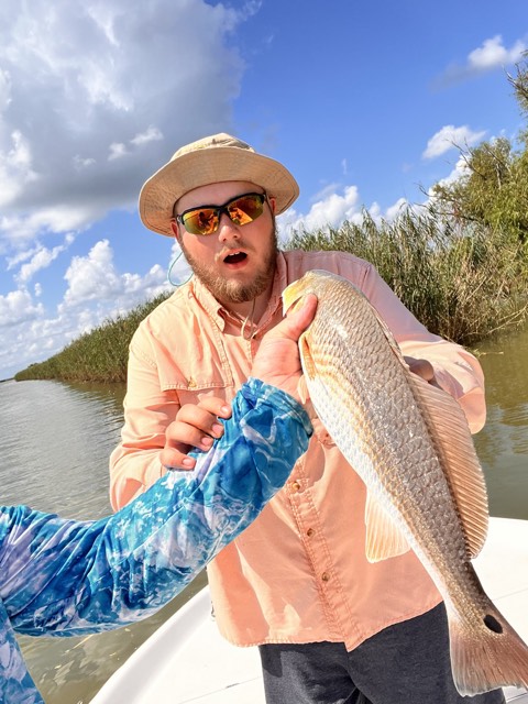 Man on boat holding up a red fish, surprised expression. Sunny day, water, green reeds.