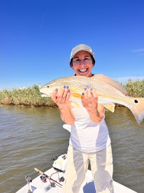 Woman in a boat holding a large redfish, smiling, against a sunny, blue sky.