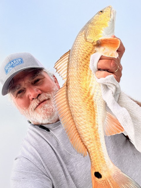 Man holding up a reddish-orange redfish with a single black spot near its tail; outdoors.
