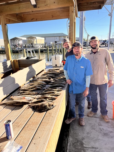 Three people pose with a large catch of fish on a dock under a covered area. Sunny day.