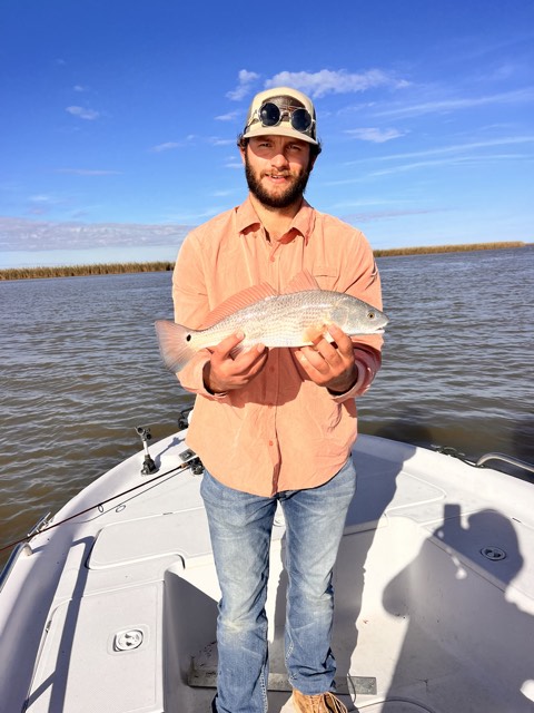 Man on a boat holds a redfish. He's wearing a hat, orange shirt, and jeans. Water and marsh in background.