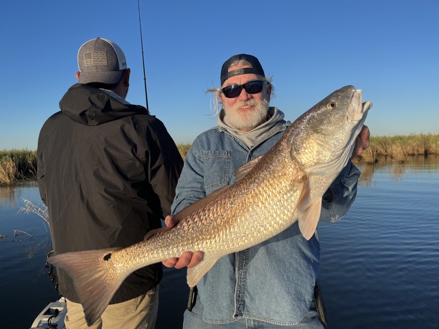 Man holding large redfish on a boat, other man behind him, blue water, sunny day.