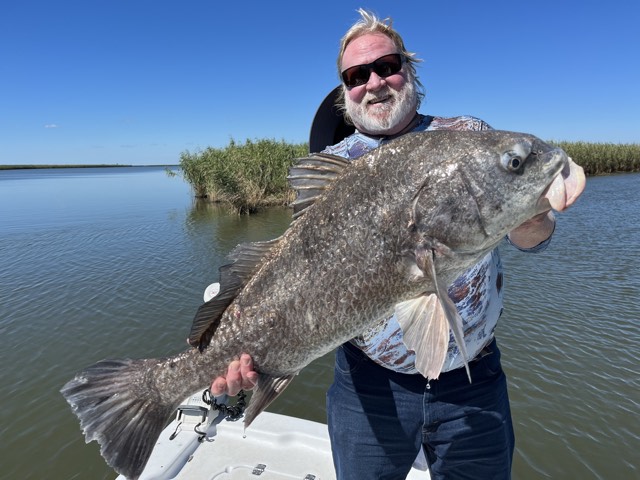 Man on boat holding large, dark fish; sunny day, water and marsh in background.
