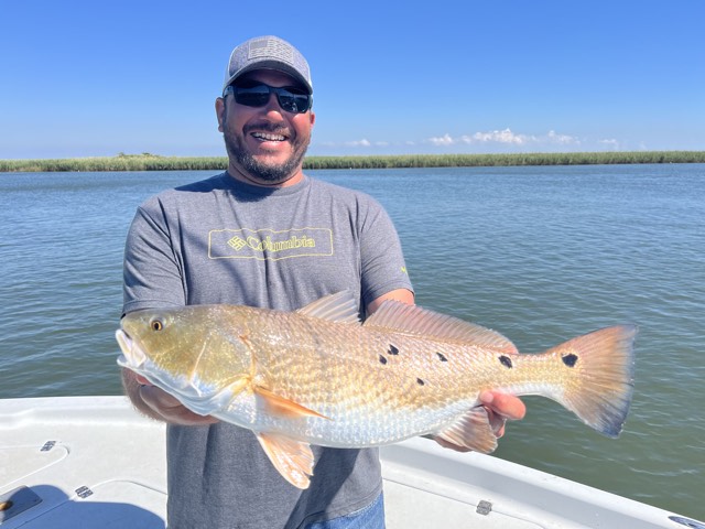 Man holding a large redfish on a boat, smiling, with water and blue sky in the background.