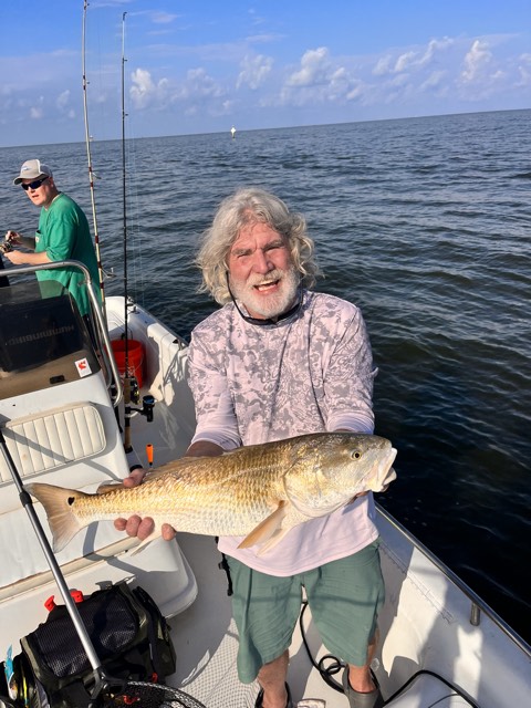Man on boat holding large fish, smiling. Another person fishing in the background. Ocean setting.