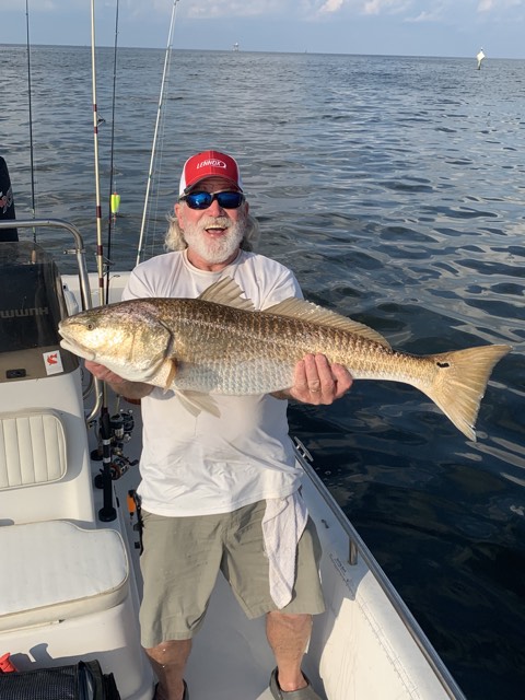 Man on a boat holding a large redfish, smiling. Ocean setting, bright sunlight.